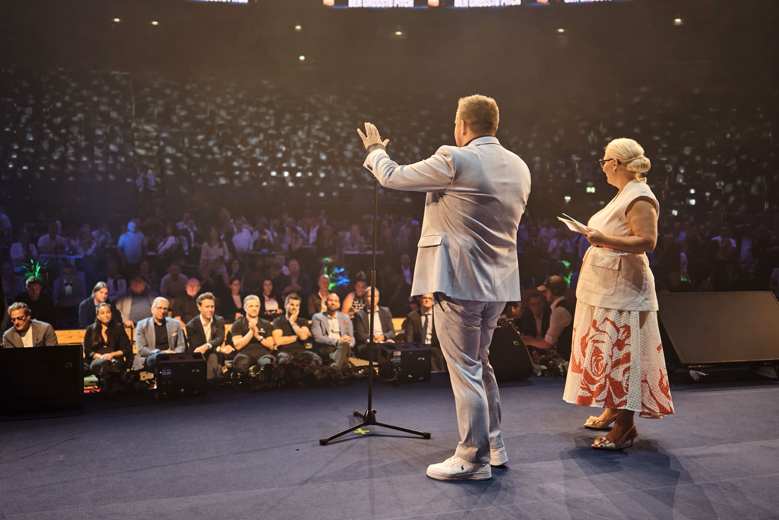 Moderationsduo auf der Bühne der LANXESS arena vor dem vollen Innenraum.