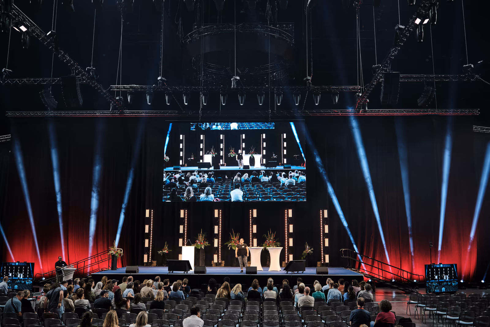 Personalversammlung in der LANXESS arena: die illuminierte Show-Bühne mit Rednerin und großer LED-Wand, davor das sitzende Publikum in blau-rotem Bühnenlicht.