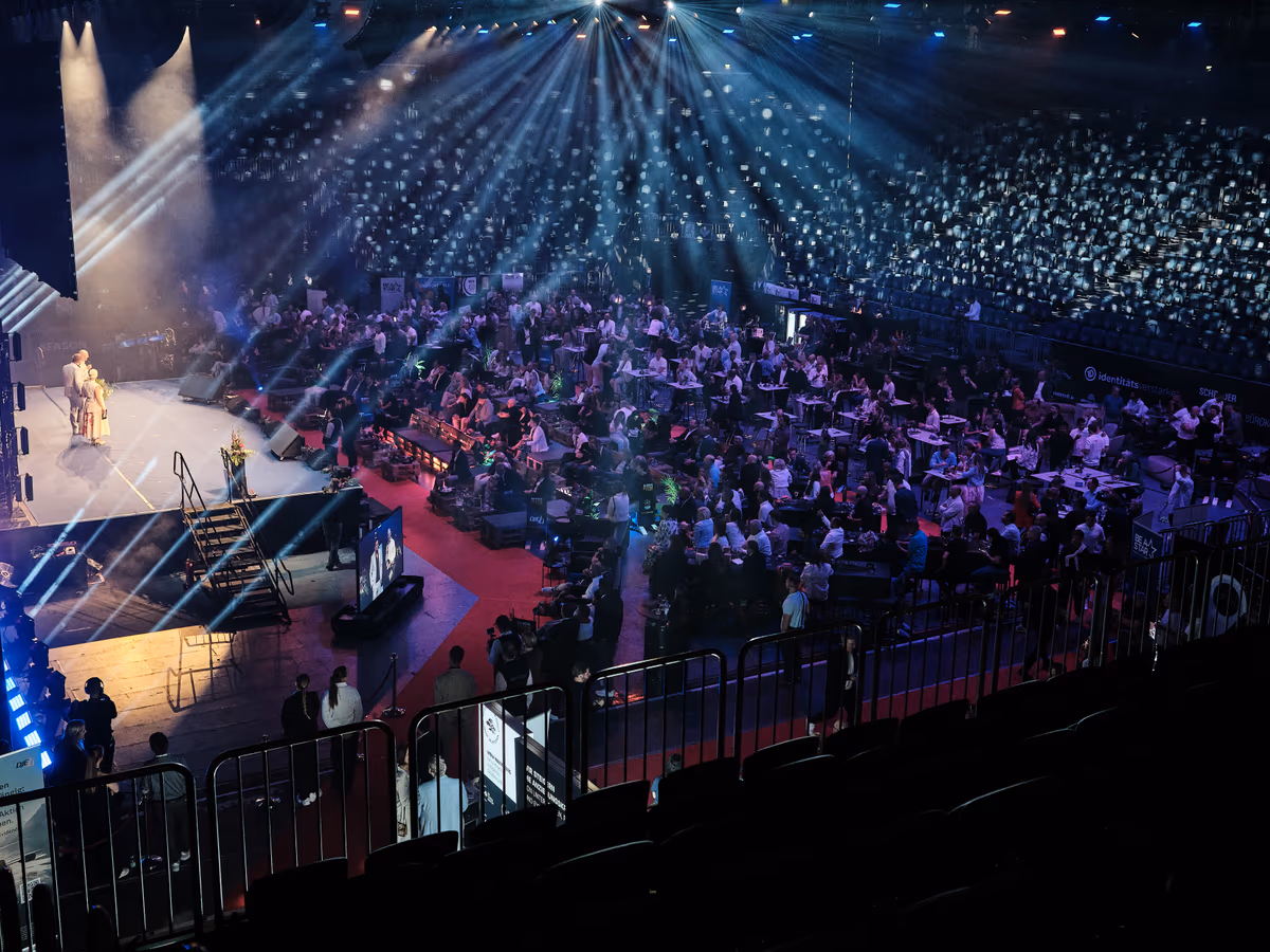 Blick von oben in die volle LANXESS arena beim größten Pitch: die beleuchtete Bühne mit Rednerin links, davor rund 1.000 Gäste an Tischen im blauen Scheinwerferlicht.