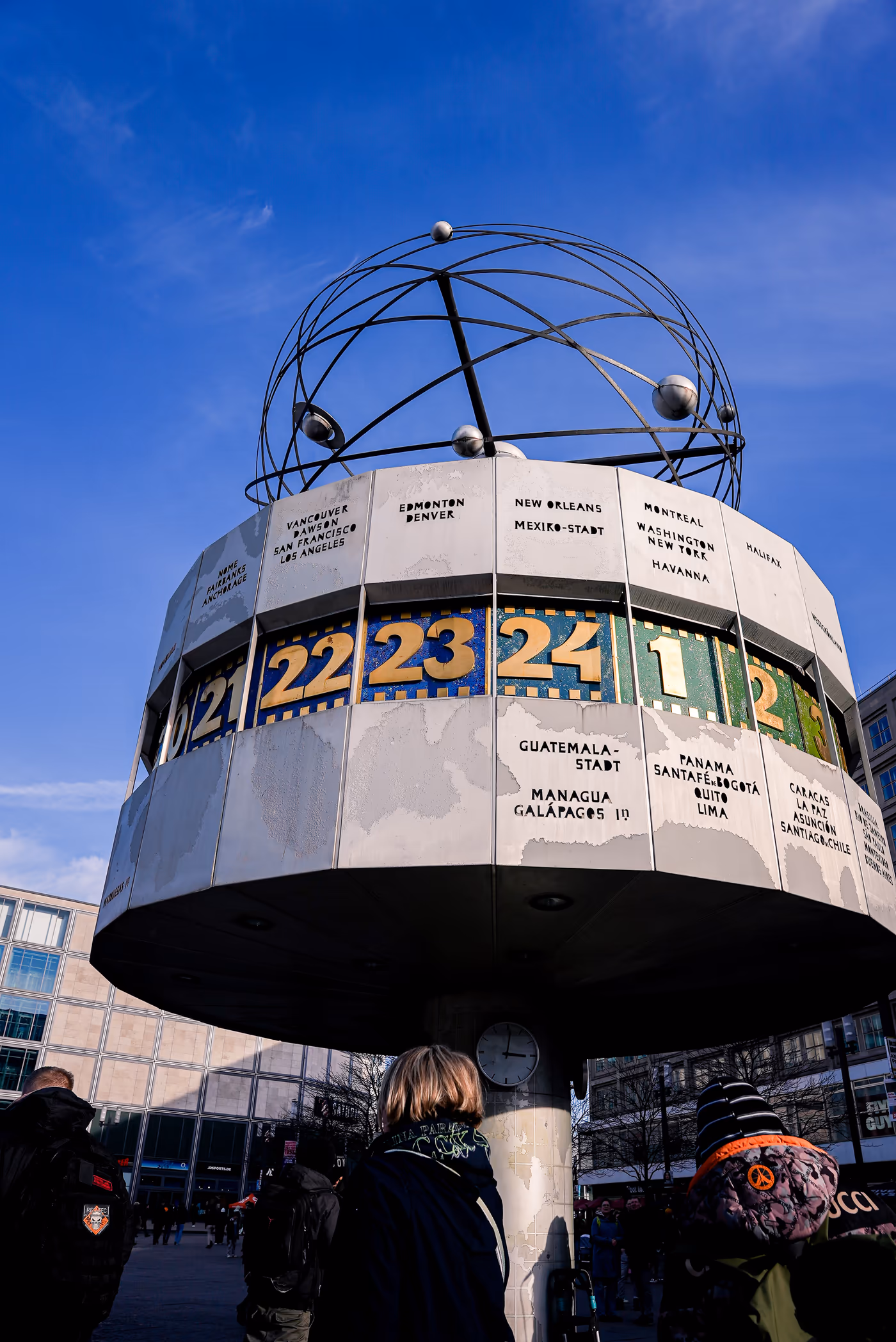 Die Weltzeituhr am Berliner Alexanderplatz vor blauem Himmel, Passanten davor.