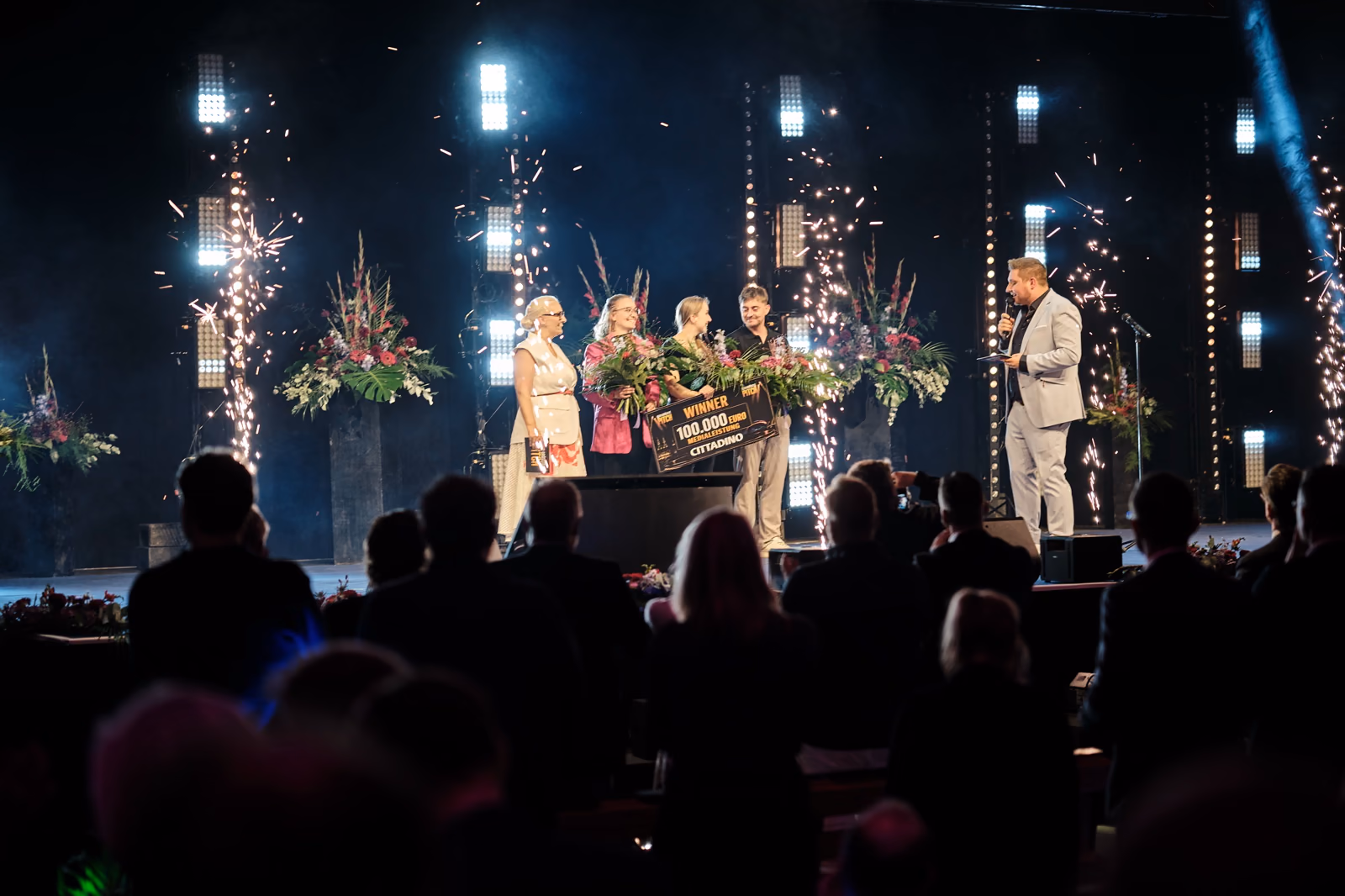 Siegerehrung auf der Bühne: das Gewinnerteam mit dem Scheck über 100.000 Euro zwischen Funkenfontänen.