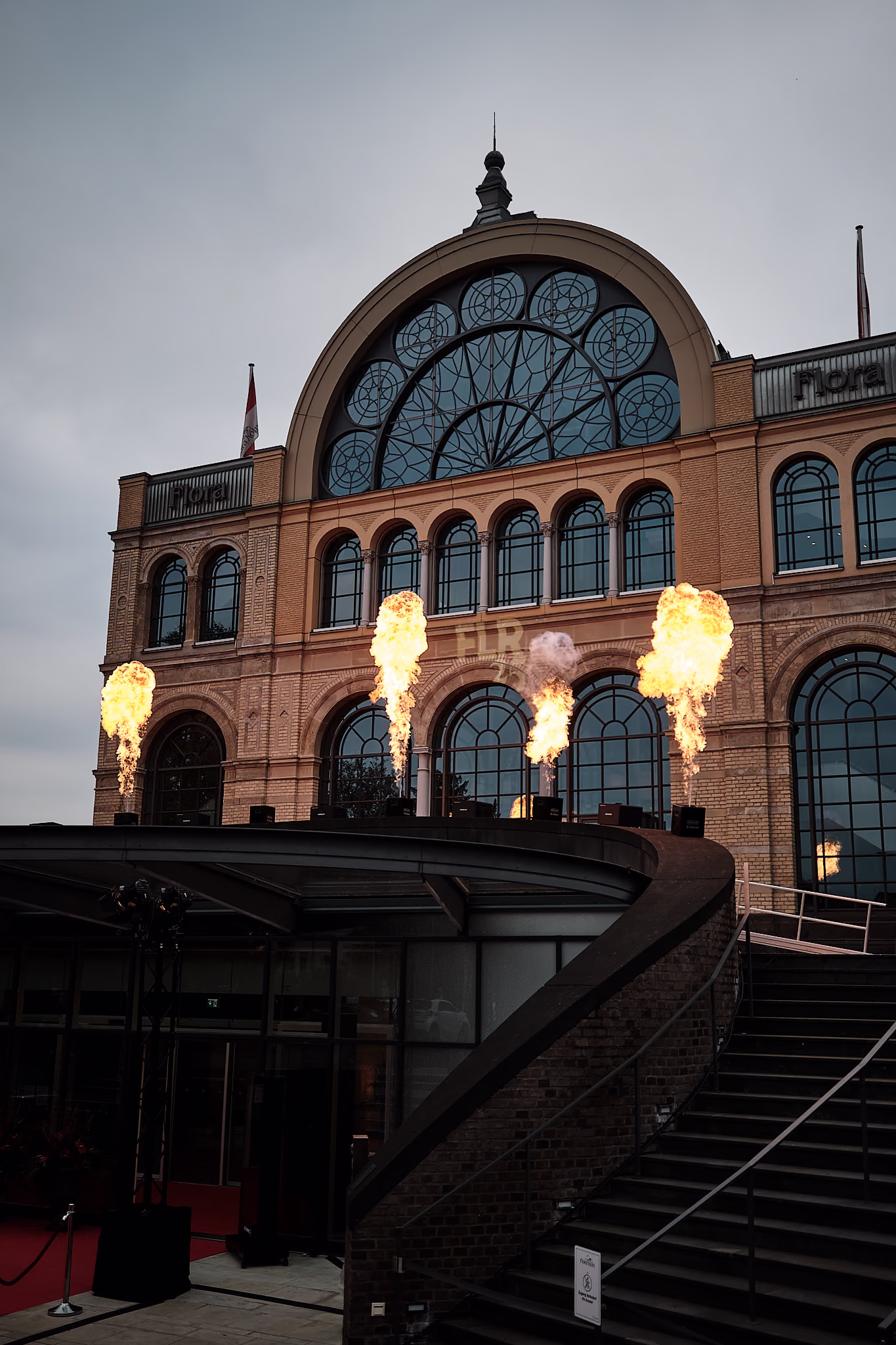 Die Flora bei Nacht, vor der beleuchteten Fassade brennen Feuersäulen.
