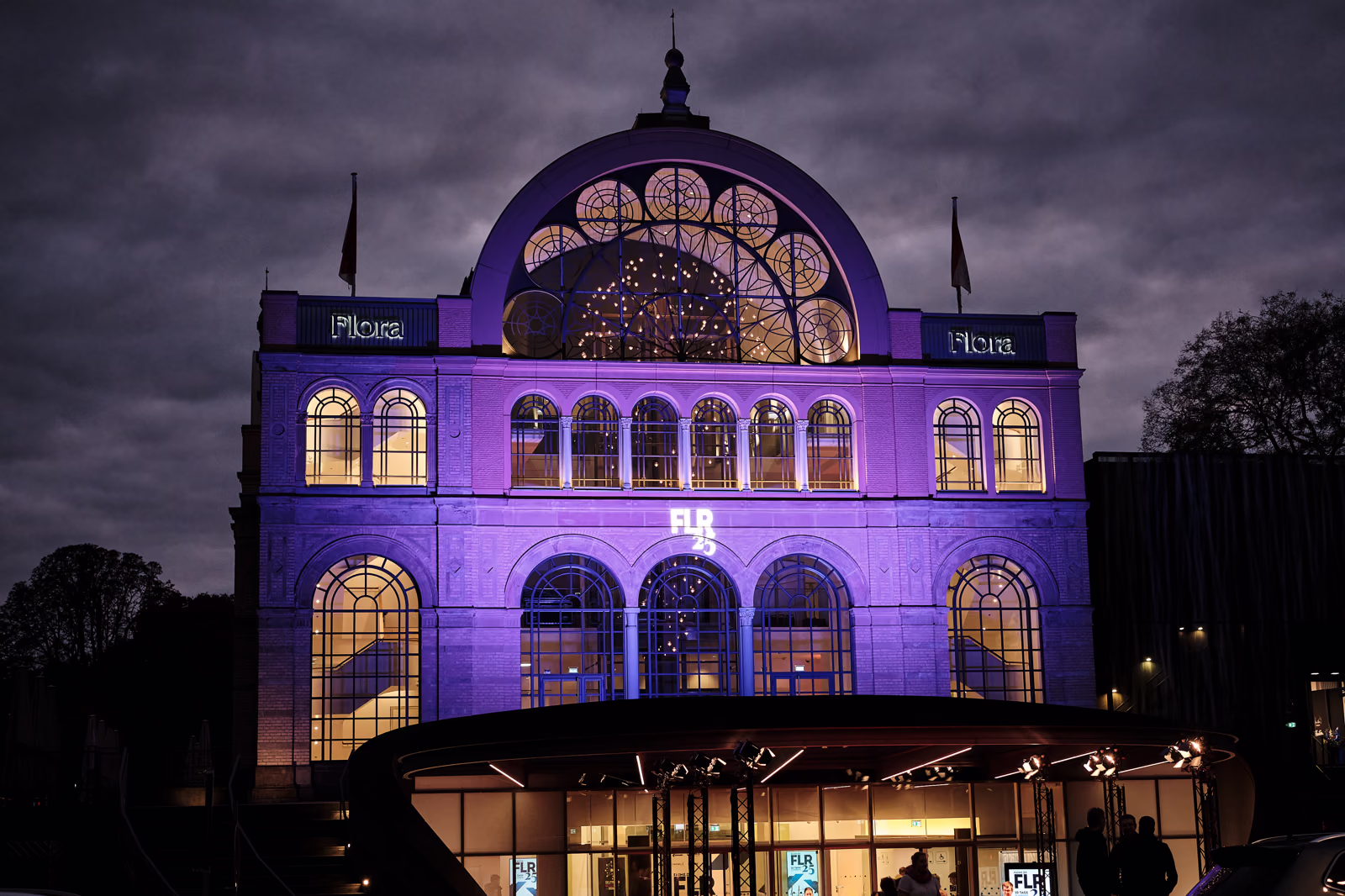 Die Flora Köln bei Nacht, in violettes Licht getaucht, hinter den Bogenfenstern leuchtet der Saal.